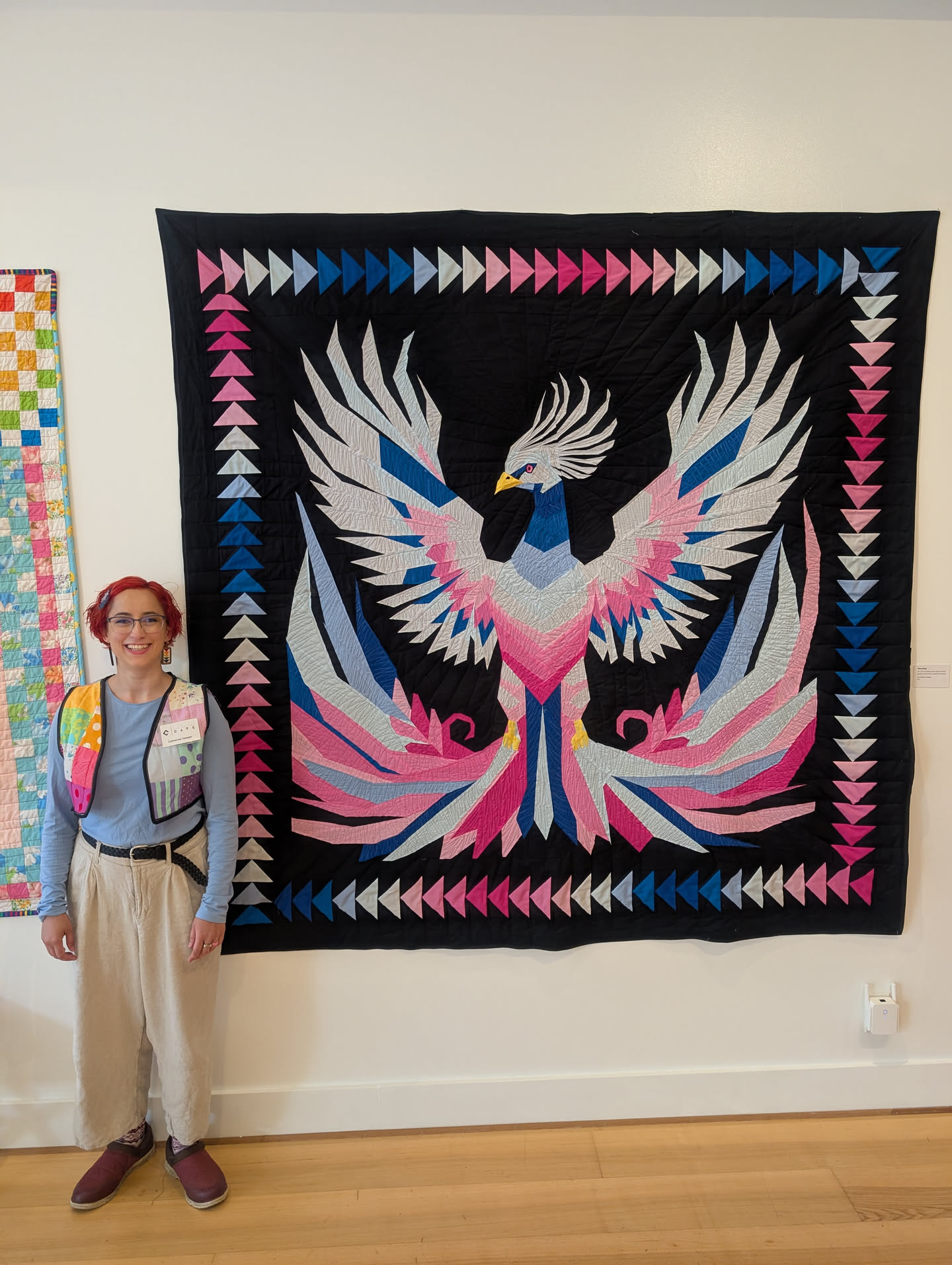 A photo of Cat standing next to her finished Becoming quilt, facing the camera and smiling.